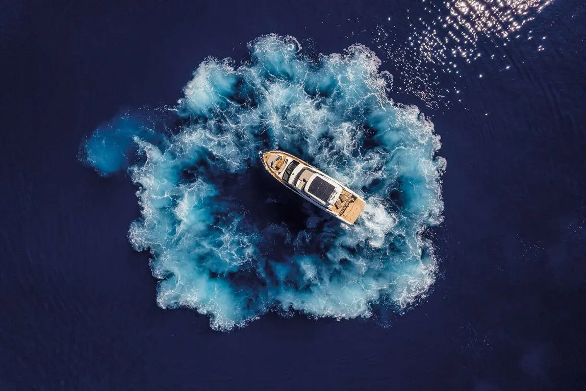 Aerial view of a yacht creating a blue spray in dark water, with sunlight reflecting on the surface.
